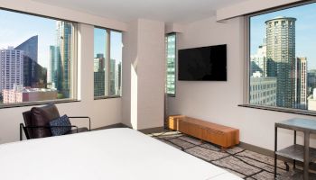 The image shows a modern hotel room with large windows offering a cityscape view, a flat-screen TV, and minimalistic furniture.