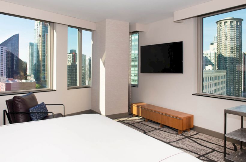 The image shows a modern hotel room with large windows offering a cityscape view, a flat-screen TV, and minimalistic furniture.
