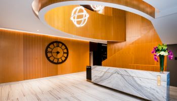 This image shows a modern lobby with a marble reception desk, wooden paneling, a large wall clock, spiral staircase, and decorative lighting.