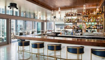 A modern bar with a wooden counter, high-back stools, and a fully stocked liquor shelf, overlooking a cityscape through large windows ending the sentence.