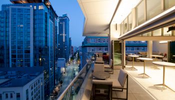 A modern cityscape at dusk featuring high-rise buildings and a stylish rooftop terrace with seating and tables. The skyline is beautiful.