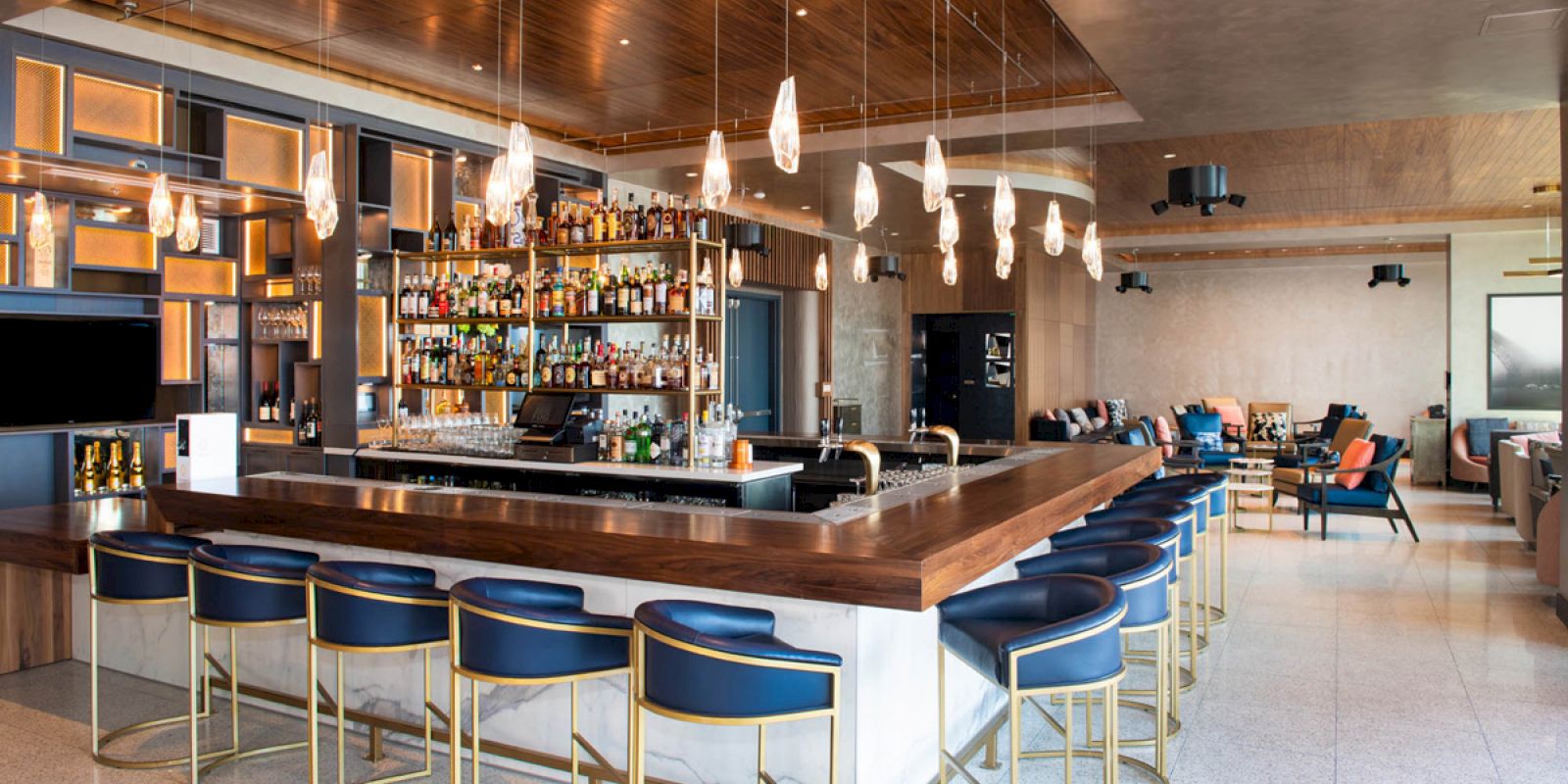 This image shows a modern, stylish bar with blue chairs, a well-stocked back bar, hanging lights, and a spacious seating area with contemporary decor.