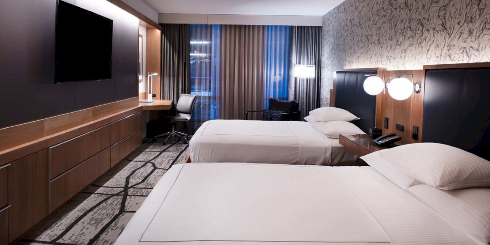A modern hotel room featuring two neatly made beds, a TV on the wall, a desk with a lamp and chair, and large windows with curtains.