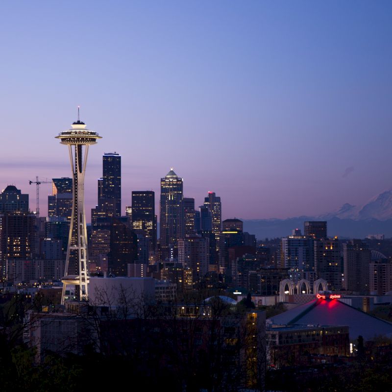 The image shows the Seattle skyline, featuring the Space Needle, lit buildings at dusk, and Mount Rainier in the background, with a clear evening sky.