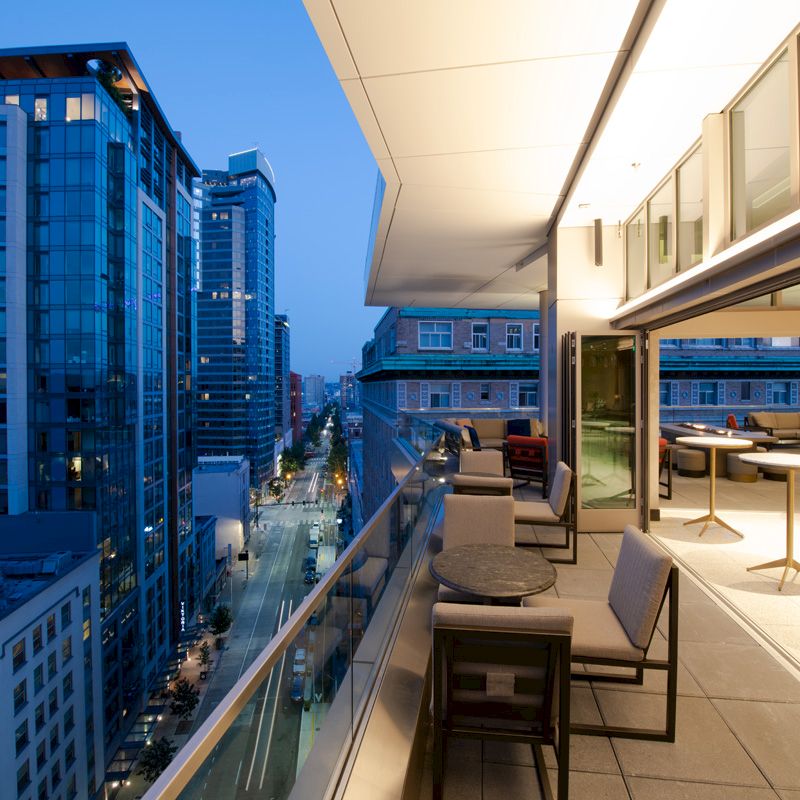 A modern rooftop terrace during evening with seating, tables, and glass railings, overlooking a cityscape with tall buildings.