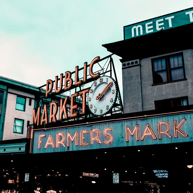This image shows a neon sign for a public market and farmers market on a building with clock, in an urban setting with other buildings in the background.