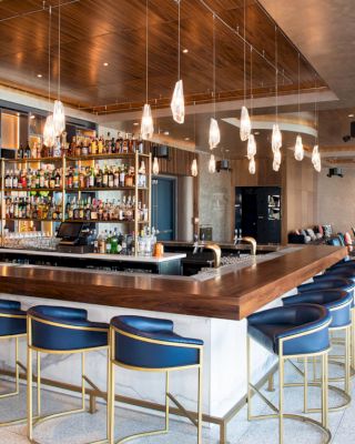 A modern bar with blue chairs, a wooden counter, pendant lights, and shelves filled with liquor bottles, located in a spacious lounge area.