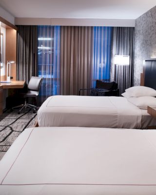 The image shows a modern hotel room with two beds, a flat-screen TV, a desk with a chair, and contemporary decor and lighting.