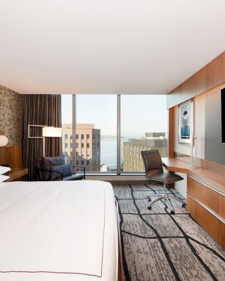 A modern hotel room with a large bed, flat-screen TV, desk, chair, and floor-to-ceiling windows offering a cityscape view.
