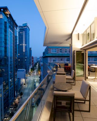 A modern rooftop balcony at dusk, overlooking a city street and tall buildings. The terrace has seating areas and open sliding doors, creating a cozy vibe.