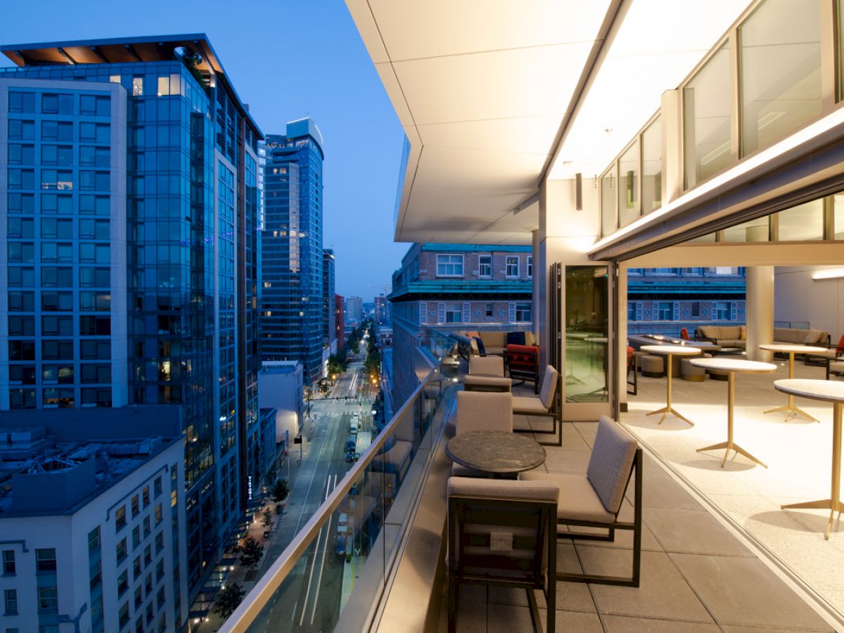 A modern balcony overlooks a cityscape with tall buildings at dusk. The balcony features outdoor seating and tables with a view of the street below.