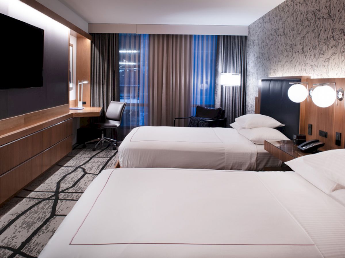 A modern hotel room with two beds, a TV on the wall, a desk by the window, nightstands with lamps, and stylish decor.