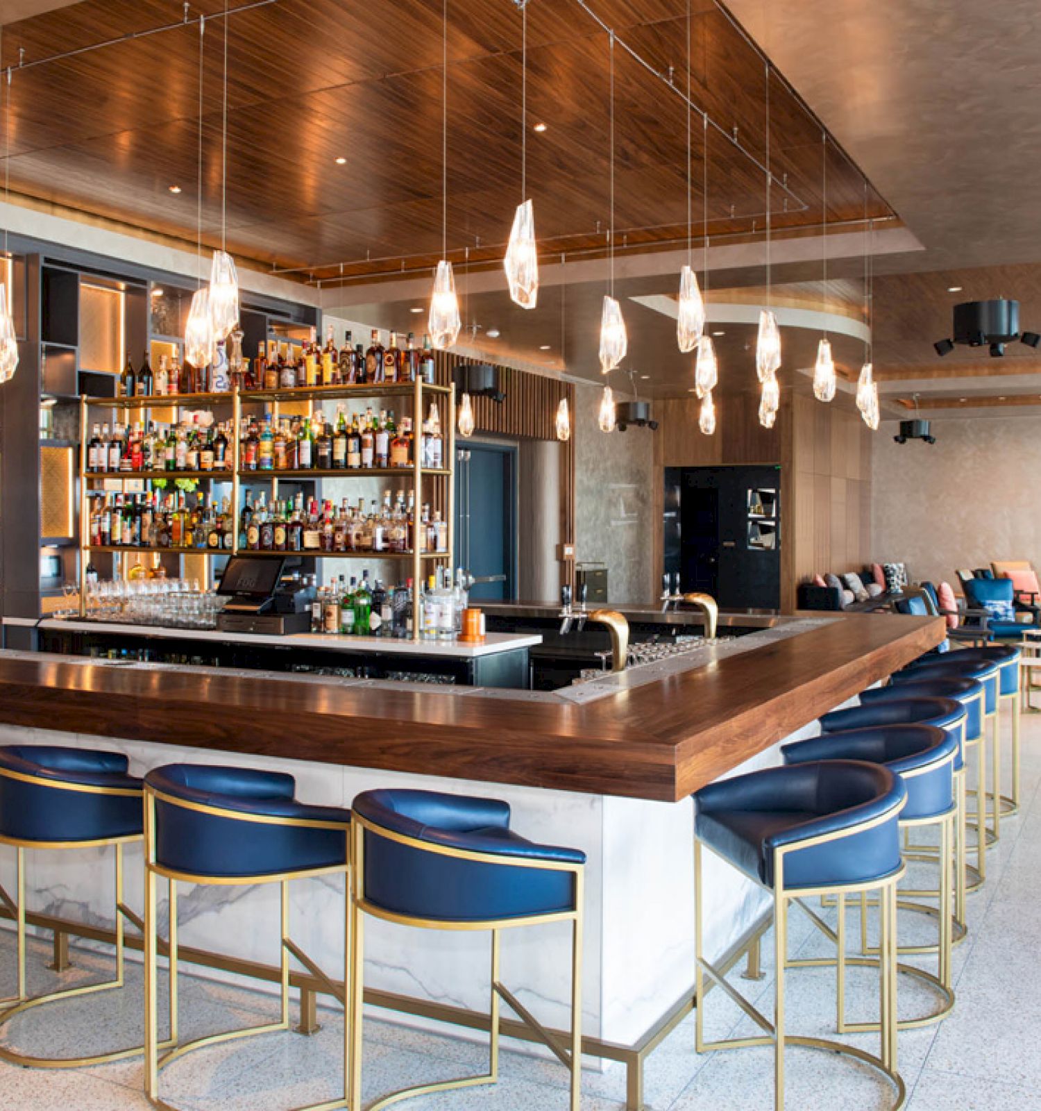 The image shows a modern bar with a wooden countertop, blue barstools, hanging light fixtures, and shelves stocked with various bottles.
