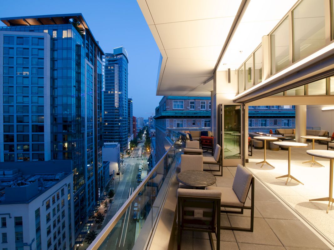 A modern cityscape at dusk, viewed from a sleek balcony with tables and chairs, overlooking a busy street and high-rise buildings.