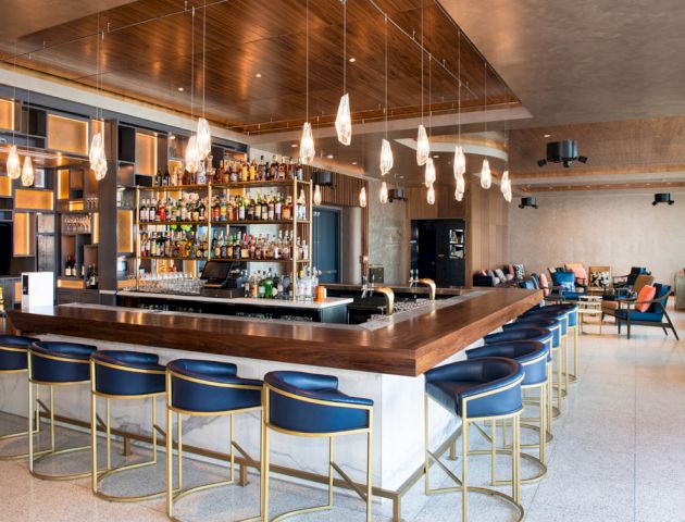 A modern bar with a wooden finish, blue cushioned stools, hanging lights, and a fully stocked liquor shelf, adjacent to a lounge area with sofas.