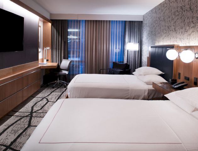 A hotel room with two beds, modern decor, a wall-mounted TV, a work desk, and warm lighting.