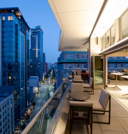 A modern balcony with tables and chairs overlooking a city street lined with tall buildings, illuminated during twilight hours.