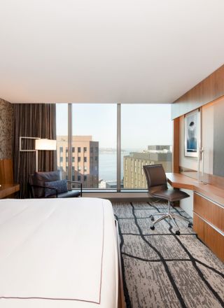 A modern hotel room with a large bed, wall-mounted TV, desk, chair, and a window with a cityscape view and water in the distance.