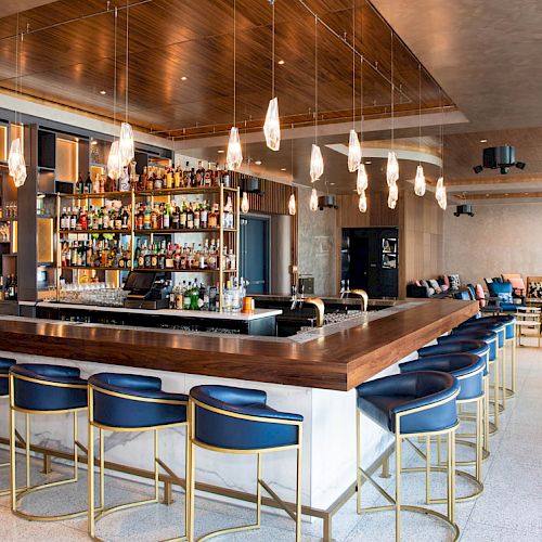 A modern, stylish bar with blue chairs, wooden accents, hanging light fixtures, and a well-stocked shelf in a spacious, upscale interior.