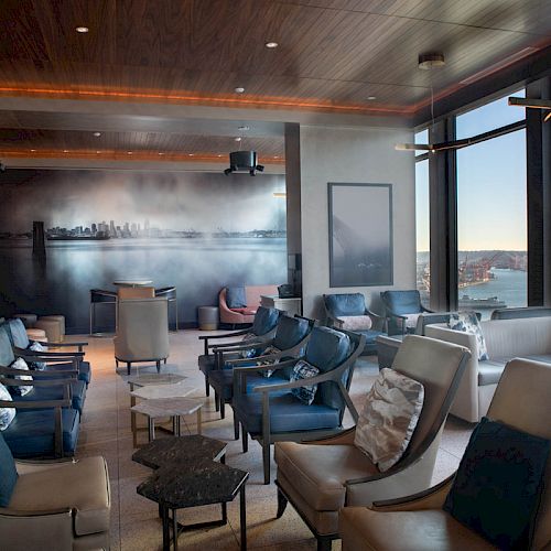 A modern lounge area with several cushioned chairs and tables, featuring large windows with an ocean view and a city mural on the back wall.