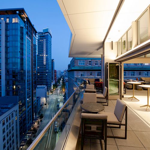 A modern cityscape at dusk, viewed from a balcony with tables and chairs. A tall building is illuminated against the evening sky.