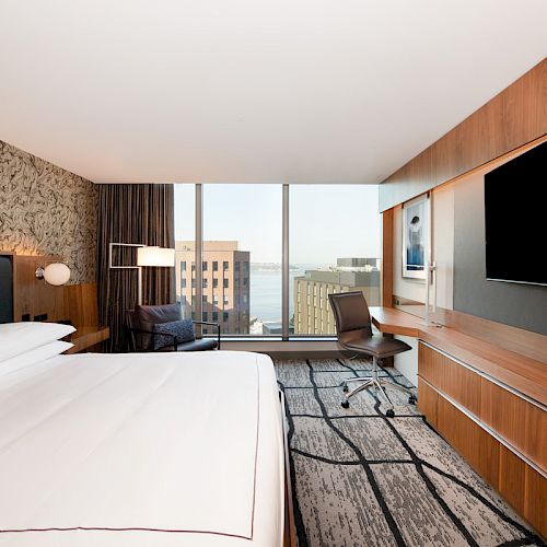 A modern hotel room with a large bed, wall-mounted TV, desk, and chair. The window offers a view of city buildings and water in the distance.