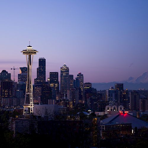 Image of Seattle’s skyline at dusk, featuring the Space Needle, downtown buildings, and Mount Rainier in the background, all under a purple sky.