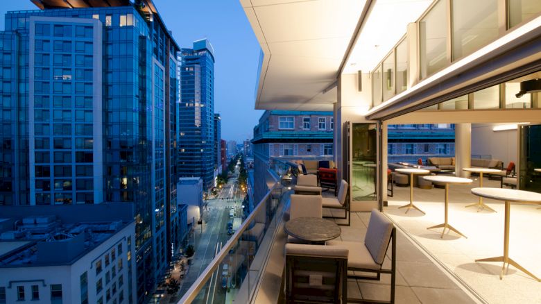 A modern balcony with seating and tables overlooks a cityscape with tall buildings at dusk, offering a mix of indoor and outdoor space.