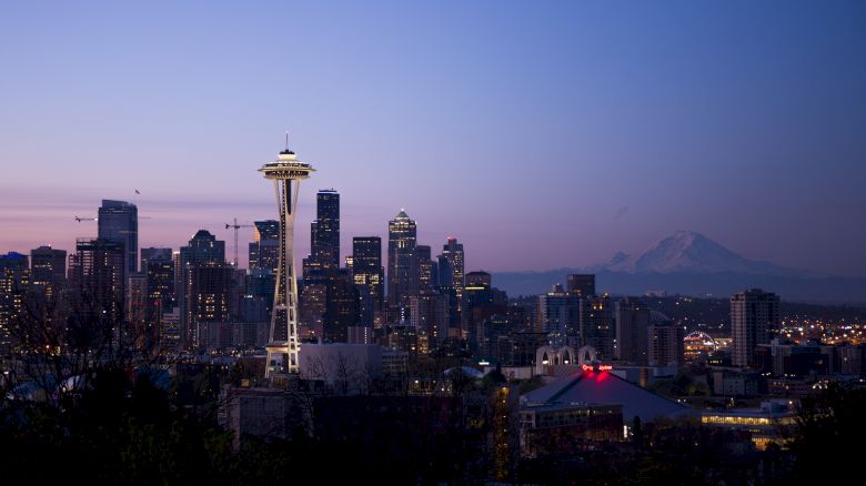 A twilight image of Seattle's skyline showcasing the Space Needle, with Mount Rainier visible in the background, and city lights beginning to illuminate.