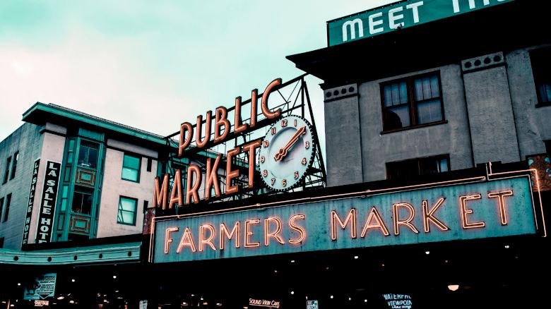 Image shows a neon sign for a Public Market and Farmers Market with a clock on a building. Other buildings and signs are visible in the background.
