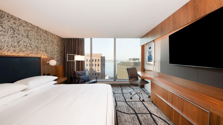 This image shows a modern hotel room with a large bed, flat-screen TV, desk, chair, and a window offering a cityscape view.