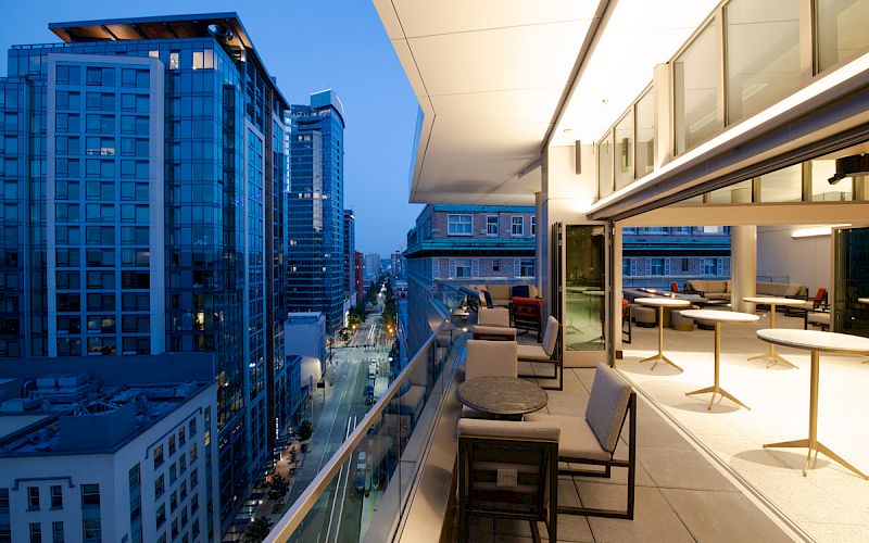 A modern urban balcony with seating overlooks a cityscape of high-rise buildings at dusk, featuring large windows and an open indoor space.