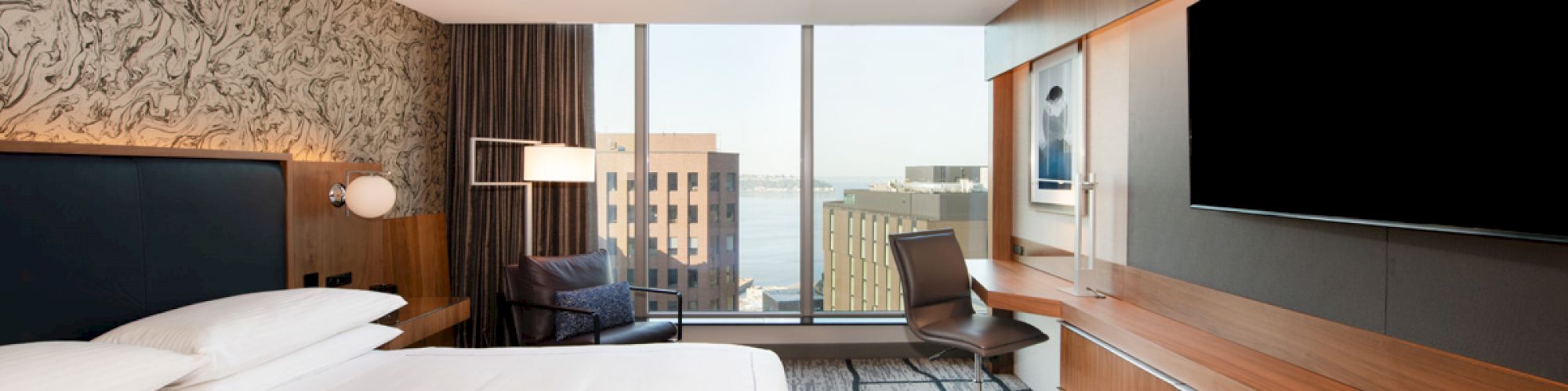 A modern hotel room featuring a large bed, desk, chair, wall-mounted TV, and a window with a cityscape view in the background.
