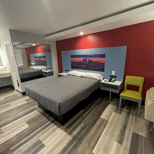 A modern hotel room with a double bed, framed cityscape artwork, a green chair, bedside tables, a sink area, and laminate flooring.