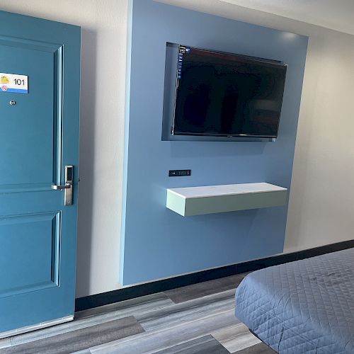 The image shows a hotel room with a bed, a wall-mounted TV, a floating shelf, and a door labeled 