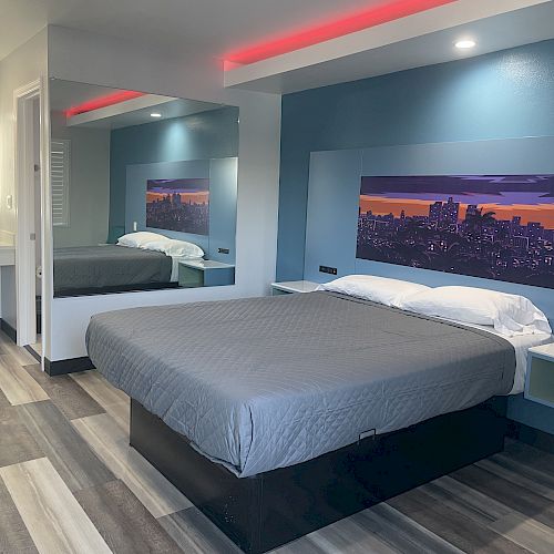 A modern hotel room with a bed, bedside tables, a painting above the bed, mirror, and a sink area with a lit mirror in the corner.