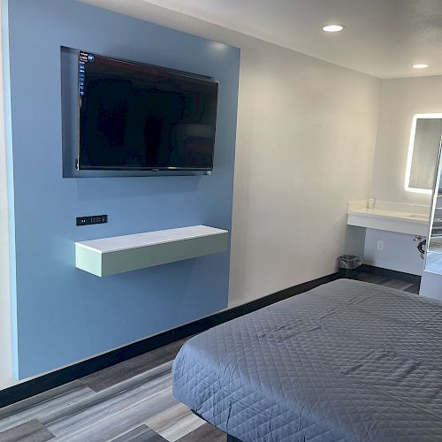 The image shows a modern hotel room with a mounted TV, a minimalist floating shelf, a bed with gray bedding, and a vanity area with a backlit mirror.