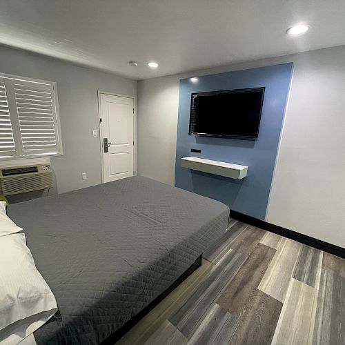 A modern, minimalist bedroom with a neatly made bed, wall-mounted TV, air conditioner, and laminate flooring. The room has a calm ambiance.