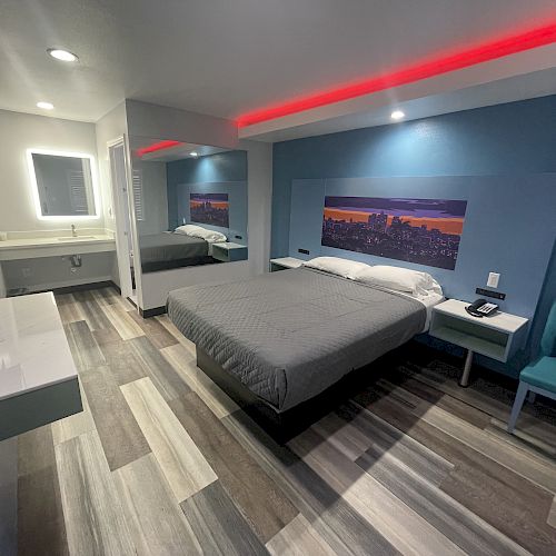 A modern hotel room with a double bed, large mirror, wall art, side table, teal chair, wall-mounted TV, and a well-lit bathroom with a standing sink.