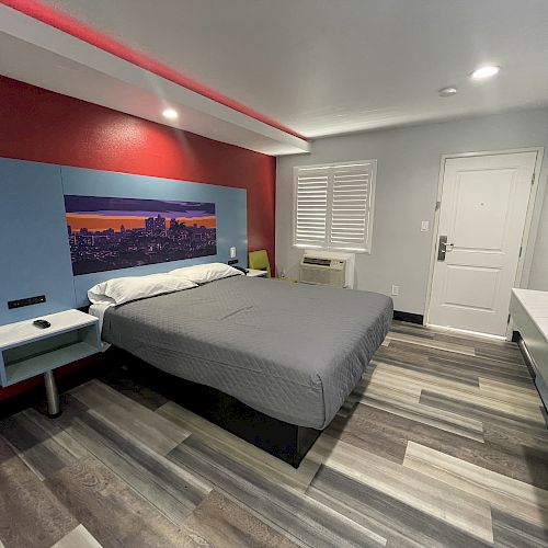 A modern hotel room with a large bed, colorful wall art, and minimalist furniture. The room has wood-inspired flooring and ample lighting.