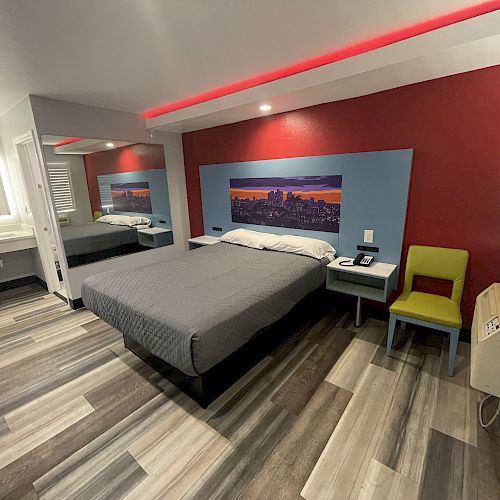 The image depicts a modern hotel room with a queen-sized bed, red and blue accent walls, a wall painting, a mirror, a green chair, and a bathroom area.