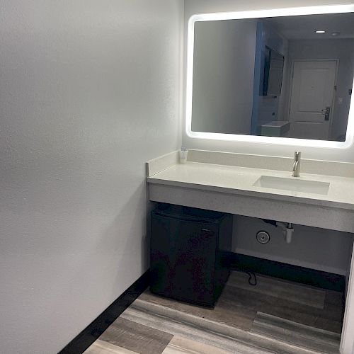 The image shows a bathroom vanity with a lighted mirror, a sink, and a waste bin underneath. The floors are tiled.