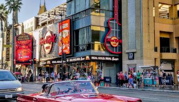 A red classic convertible car drives down a bustling street lined with iconic shops, theaters, and a giant guitar sign, possibly in Hollywood.