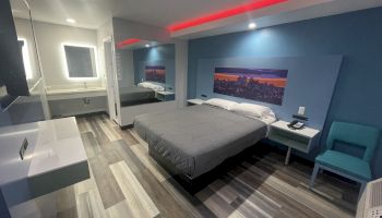 A modern hotel room features a bed, a nightstand, a chair, a wall-mounted flat-screen TV, a cityscape picture, and a bathroom area with a backlit mirror.