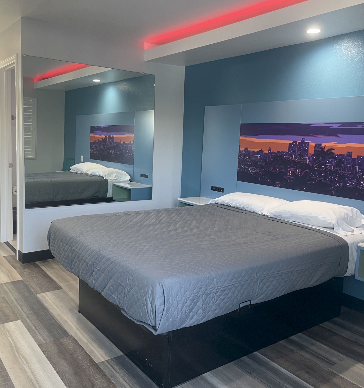 A modern hotel room with a bed, cityscape artwork, a wall mirror, and a small counter area with a sink and lighting on the left side of the image.