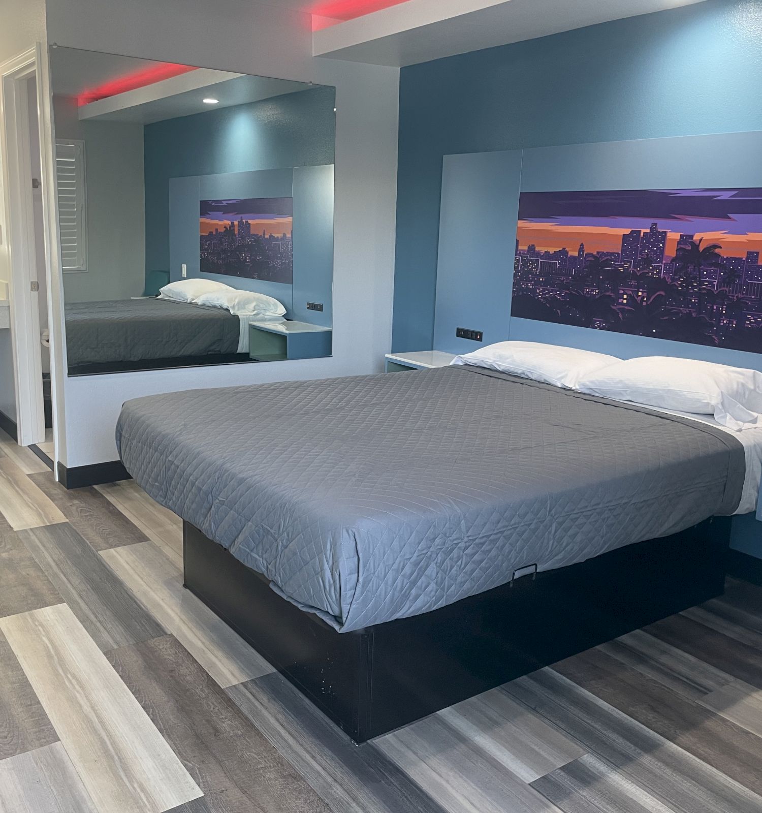 A modern hotel room with a large bed, gray bedspread, artwork above the bed, a mirror, a desk, and a bathroom sink visible in the corner.