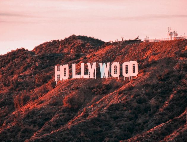 The image shows the famous Hollywood sign situated on a hill with a backdrop of greenery and a pink-hued sky, taken at sunset.