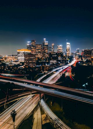 A nighttime cityscape featuring illuminated skyscrapers and busy highways with streaks of lights trailing from moving vehicles.