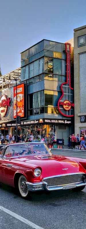 A vintage red convertible drives down a busy city street lined with colorful storefronts and neon signs, with people walking on the sidewalk.