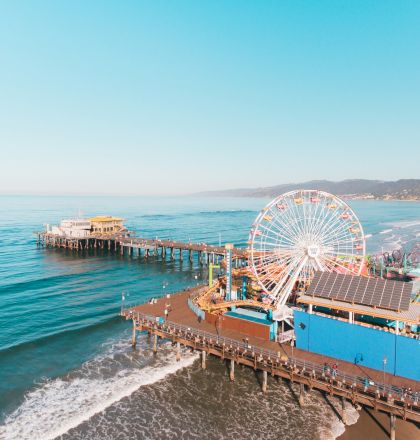 An amusement park with a Ferris wheel is on a pier extending into the ocean, with a sandy beach and distant mountains in the background.